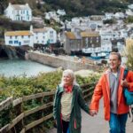 A senior man and his wife holding hands walking up a hill on a footpath looking away from the camera at the view. The fishing village of Polperro is behind them.