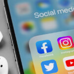 showing social media apps icons of Facebook, Instagram, WhatsApp, Twitter, Youtube, TikTok - on screen smartphone iPhone with AirPods closeup.