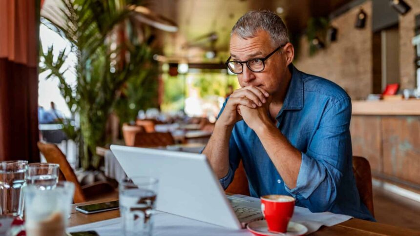 Middle-aged white man wearing glasses, staring into space over the top of his laptop in a coffee shop