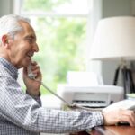 Smiling senior white man talking through telephone while using laptop at desk.