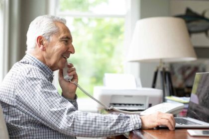 Smiling senior white man talking through telephone while using laptop at desk.