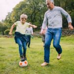 Three generation family are playing football together in a field. There are two boys, their father and their grandfather.
